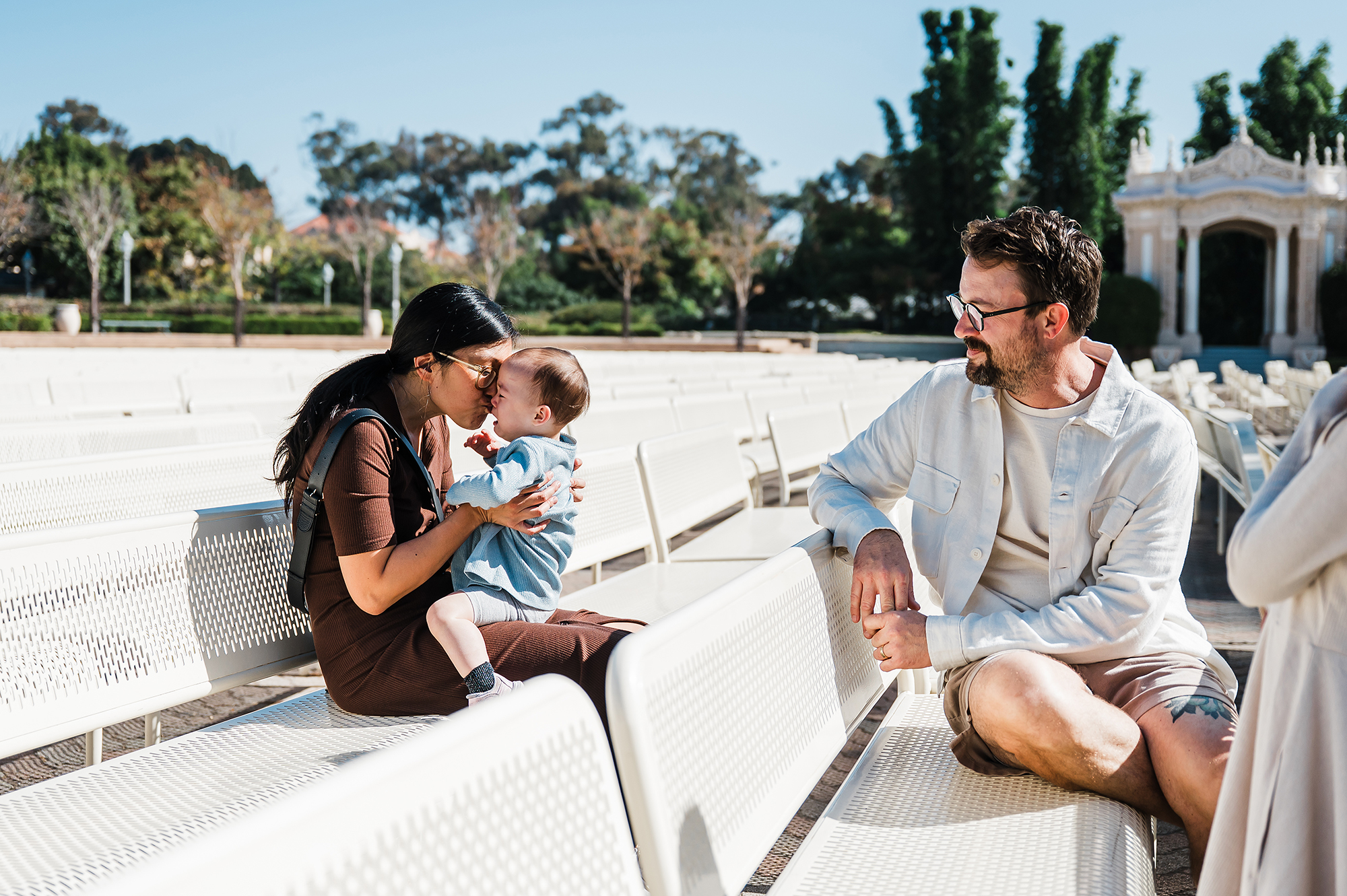 Mom comforting her baby on a bench while dad watches during a relaxed San Diego family session in Balboa Park