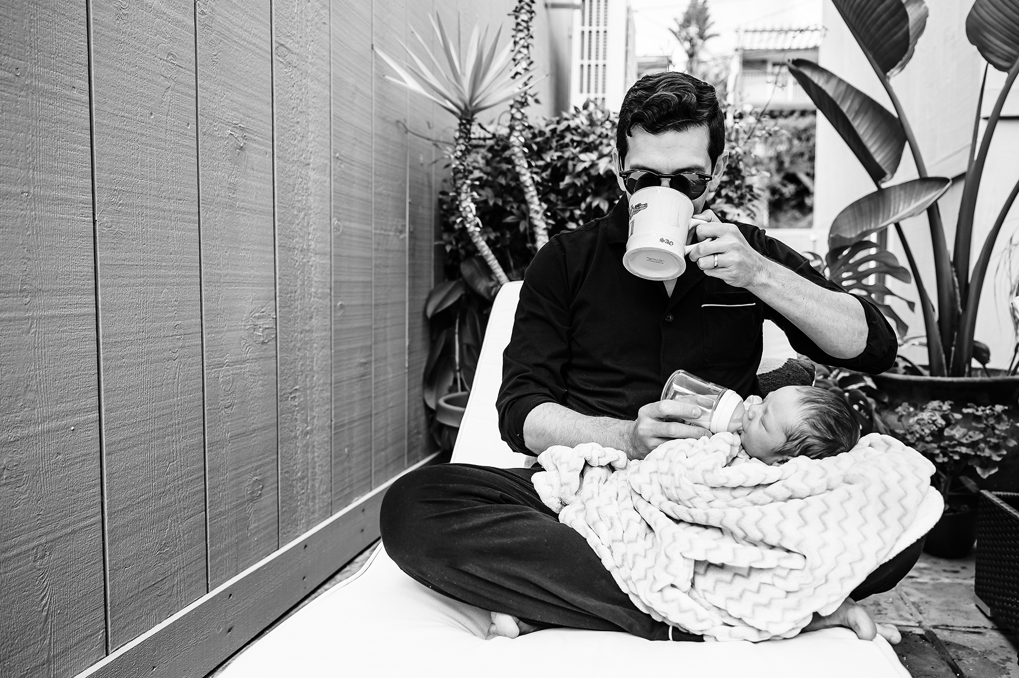 Father drinking coffee while bottle-feeding his newborn in his lap in San Diego backyard.