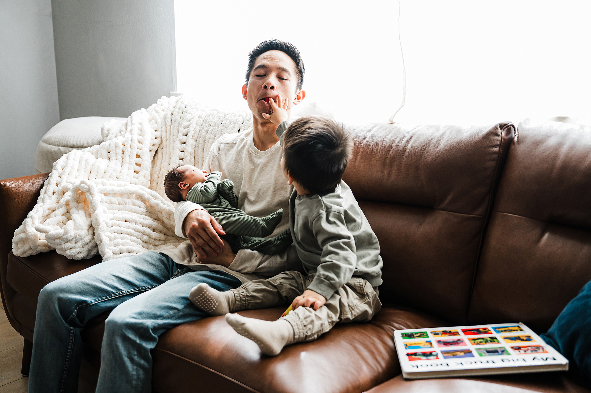 Father holding his newborn on the couch while his older child reaches up to touch his face during in home newborn photography session.
