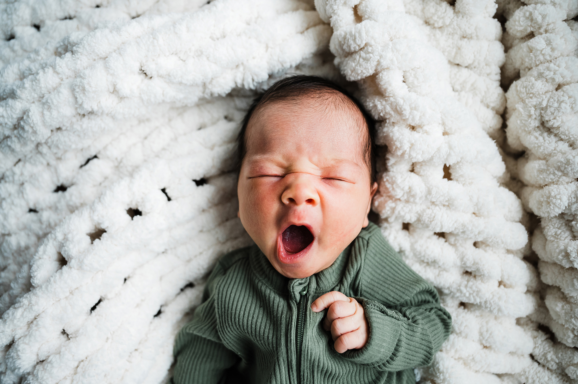 Newborn in a green outfit yawning while nestled in a white knitted blanket.
