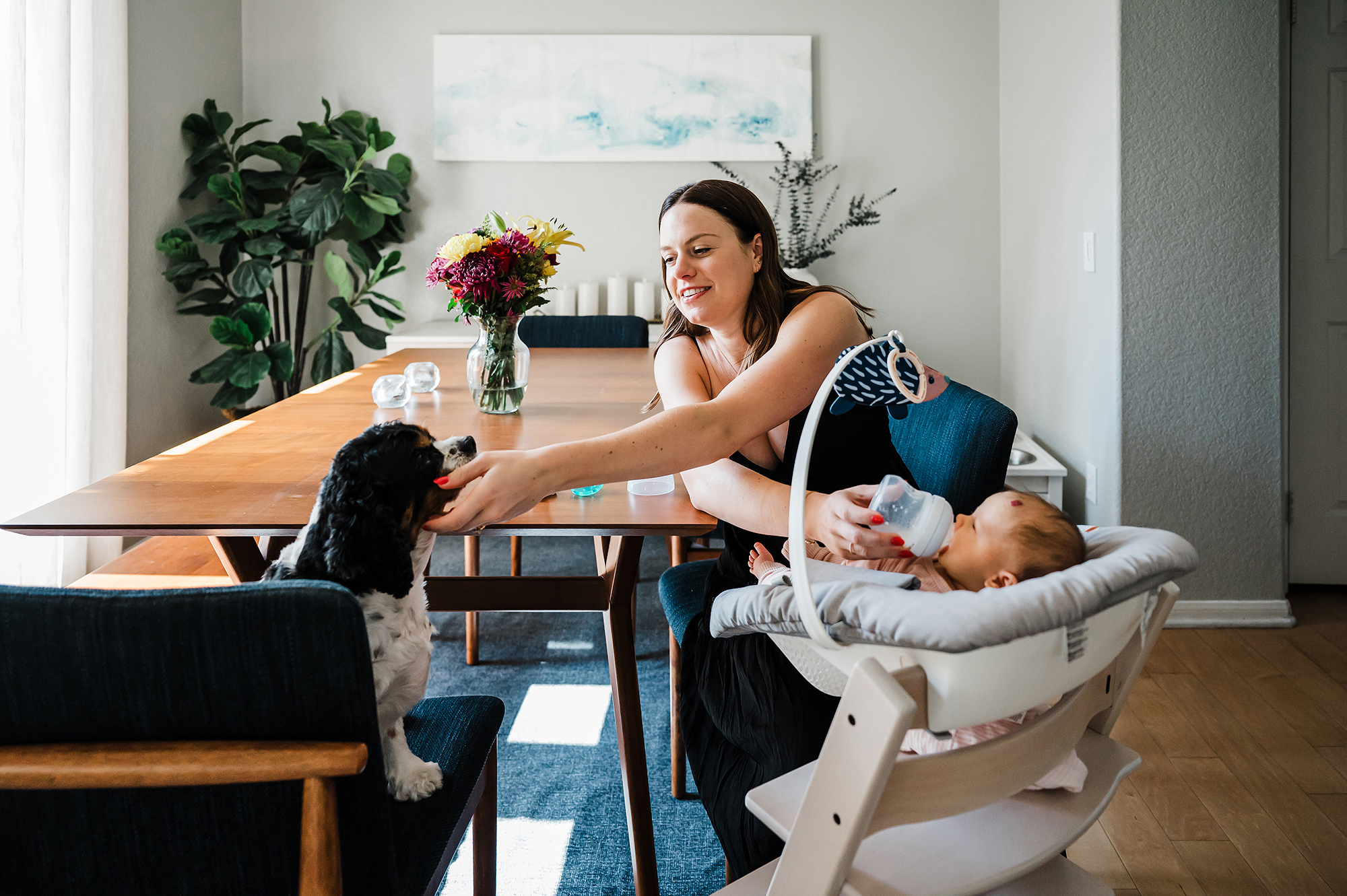 Mother bottle-feeding her newborn in a reclined baby seat while petting the dog beside her in San Diego newborn session.