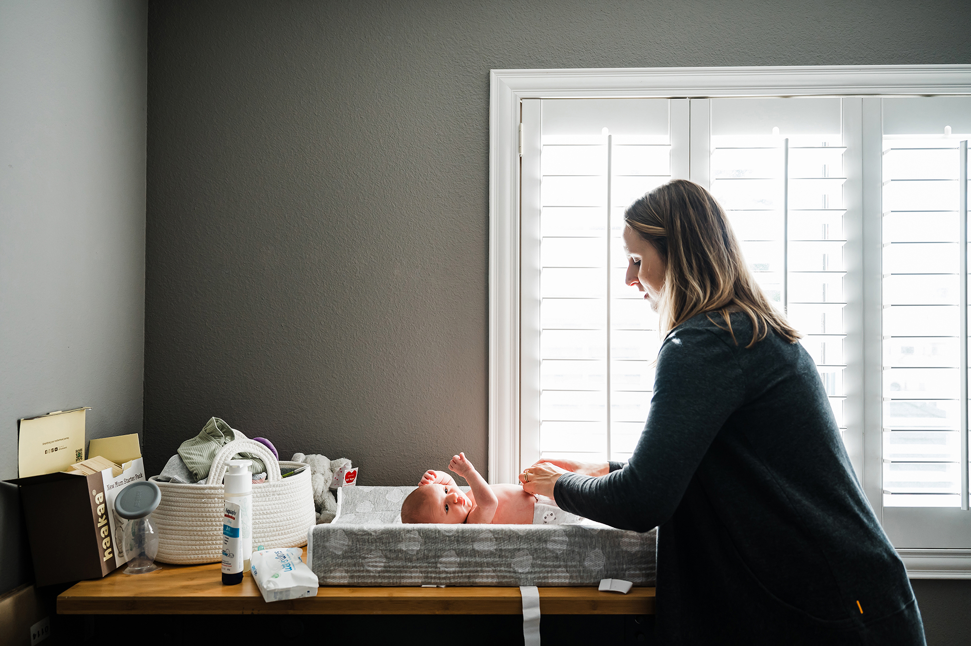 Mother changing her newborn’s diaper on a changing table beside the bedroom window.